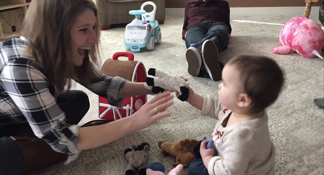 Devra playing with a toddler on the floor, holding up a hand puppet during a therapy session.